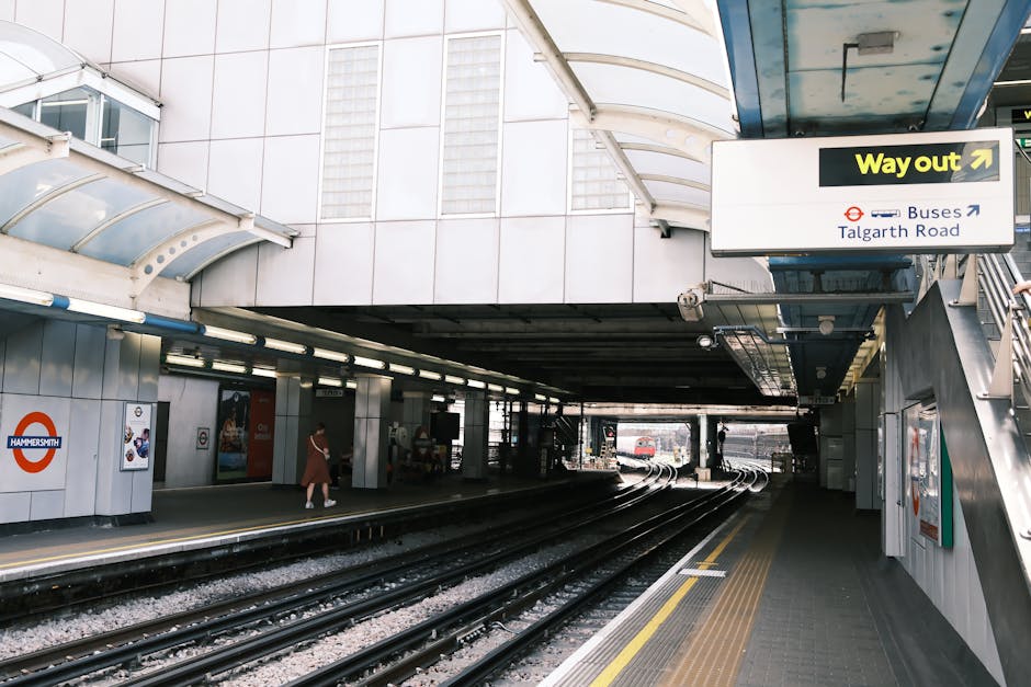 The image shows the entrance and platform area of Hammersmith station, part of the London Underground, with the station sign on the left displaying the iconic roundel logo. The platform has grey paving tiles, and train tracks extend into the distance, curving slightly to the right. There is a metal and glass canopy overhead providing shelter, with fluorescent lighting beneath. A few passengers are visible, including a woman walking along the platform with a small bag, waiting near the edge. On the right side, there is a staircase leading up, with a yellow 'Way out' sign and additional signage for buses to Talgarth Road. The station surrounding environment appears modern, clean, and well-maintained. The image highlights the typical infrastructure associated with urban transport stations, which can be relevant context for house removals involving the disassembly or transport of household items through this area, as handled by Man with Van Southfields.