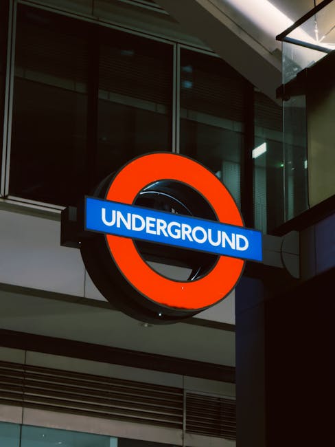 A large, circular London Underground sign featuring a bold red outer ring and a black inner circle, mounted on the exterior wall of a modern building with glass windows and metal architectural elements. A blue rectangular sign with white text reading 'UNDERGROUND' spans across the middle of the circle. The sign is positioned outdoors, illuminated by the ambient lighting of the building façade, and is part of a transportation hub often used during home relocation or furniture transport processes in Southfields. The surrounding environment includes reflections on the glass surfaces and structural details of the building, relevant to urban moving logistics and professional removals services provided by Man with Van Southfields, as seen in the context of house removals and packing operations near the Tube station.