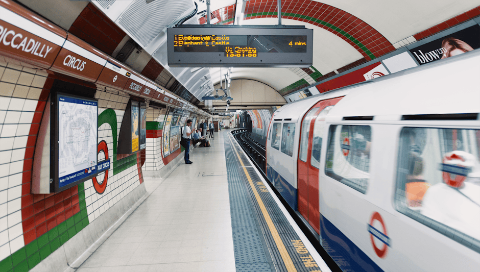 The image shows the entrance and platform area of Hammersmith station, part of the London Underground, with the station sign on the left displaying the iconic roundel logo. The platform has grey paving tiles, and train tracks extend into the distance, curving slightly to the right. There is a metal and glass canopy overhead providing shelter, with fluorescent lighting beneath. A few passengers are visible, including a woman walking along the platform with a small bag, waiting near the edge. On the right side, there is a staircase leading up, with a yellow 'Way out' sign and additional signage for buses to Talgarth Road. The station surrounding environment appears modern, clean, and well-maintained. The image highlights the typical infrastructure associated with urban transport stations, which can be relevant context for house removals involving the disassembly or transport of household items through this area, as handled by Man with Van Southfields.