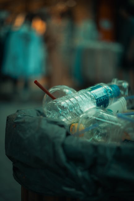 Close-up image of a large plastic bin filled with discarded empty plastic bottles and other waste materials, some wrapped in plastic film or with labels partially visible. The bottles are translucent, with one featuring a blue label and others showing clear plastic. The bin is situated outdoors near a moving or waste disposal area, with a blurred background that includes clothing or fabric and hints of a busy environment, possibly near a loading zone for home relocation or furniture transport services. The scene captures the process of disposal or waste collection associated with moving logistics, with natural lighting highlighting the plastic clutter in the bin, which sits on a flat surface such as pavement or inside a waste collection point, consistent with the context of bulky waste disposal linked to house removals in SW18. Man with Van Southfields occasionally manages such waste clearance as part of their comprehensive removal services, including furniture and bulky waste removal during home relocation procedures.