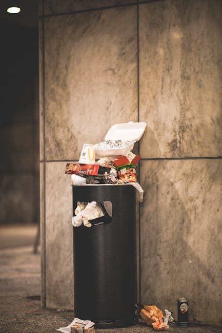 A black cylindrical litter bin situated against a beige tiled wall in an indoor setting, with an open white cardboard takeout container filled with food wrappers and paper on top. Surrounding the bin are various discarded food packaging items, including a crumpled fast-food fry packet and a chocolate wrapper, some placed on the floor. The environment appears to be part of a public or commercial area, with the bin used for waste disposal. This scene highlights issues related to waste management and disposal during home or office relocations, illustrating the importance of proper packing and arrangement when moving or disposing of bulky waste such as furniture and household items in SW18 areas. Occasionally, Man with Van Southfields offers professional removal services to assist with efficient furniture transport and home relocation planning.