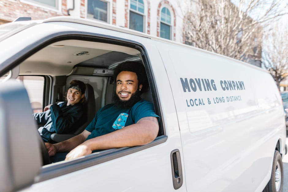 Inside a white moving van parked outside a residential building, two men are seated in the driver's and passenger's seats, smiling at the camera. The man in the driver's seat has dark curly hair and a beard, wearing a blue T-shirt, while the man in the passenger seat has dark hair with a headband and is dressed in a dark jacket. The van's side panel displays the company name 'Moving Company' along with the description 'Local & Long Distance,' indicating a professional house removal service. Visible through the open driver's side window, the interior of the vehicle includes packing materials such as cardboard boxes and protective blankets, which are typical for furniture transport and packing during a home relocation. The background shows trees and multi-storey buildings, suggesting an urban setting. The scene depicts a loading or preparation process for furniture transport and moving operations, supported by the presence of equipment like straps or trolleys often used by [COMPANY_NAME] during packing and moving tasks, particularly for a late-notice move in Southfields.