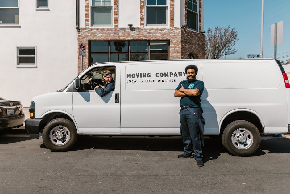 A man with long, wavy hair and a bandana is sitting in the driver's seat of a white moving van marked with the words 'MOVING COMPANY, LOCAL & LONG DISTANCE' on the side. The van is parked outside a residential building in Southfields under a clear blue sky, with modern buildings and trees visible in the background. The man is smiling at the camera, with his left arm resting on the window ledge. Inside the vehicle, visible objects include cardboard boxes and a fabric-moving blanket, which are likely packed for a home relocation. The scene captures the loading and transport process typical of professional removals, with the van positioned close to the building entrance for efficient loading of furniture and packed items. Man with Van Southfields employs these vehicles and equipment, such as straps and trolleys, to assist with home furniture transport and packing and moving services during late-notice or same-day relocations.