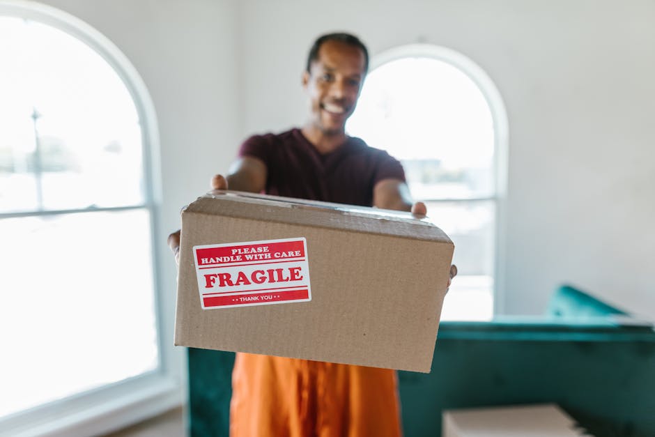 A man with Southfields-based Man with Van Southfields is inside a well-lit room, holding out a medium-sized cardboard box towards the camera during a home relocation process. The box is sealed with tape and features a red and white sticker indicating it is fragile, with the text 'Please Handle With Care, Fragile, Thank You.' The man appears to be smiling, wearing casual attire, and standing in front of a large arched window that lets in natural light, illuminating the interior space. In the background, there is furniture covered with protective blankets to prevent damage during furniture transport, and a doorway can be seen behind him. The scene captures the packing and loading aspect of a professional removals service, with attention to careful handling of delicate items during moving and relocation logistics, aligned with the services offered by [COMPANY_NAME] on their website, manwithvansouthfields.co.uk.