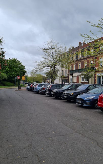 The image shows a parking area on a residential street with several parked cars along the curb, including sedans and small hatchbacks, under an overcast sky. To the right, there are multi-storey brick and stone buildings with traditional architecture, featuring windows with white frames, some with small balconies. The pavement appears slightly cracked, and there are a few trees with light foliage, indicating early spring or late autumn. In the background, there are traffic signs and a small lamp post. This scene likely depicts an urban environment suitable for house removals or furniture transport, with no vehicles blocking access to the property entrances. As part of the moving process, [COMPANY_NAME] may use such parking areas to load or unload furniture and boxes during home relocation or packing and moving operations, ensuring compliance with local parking restrictions to avoid fines, especially relevant near Southfields. The image captures a typical scene where strategic planning with parking suspensions could be necessary during scheduled removals, as highlighted in the Southfields Parking Suspensions page.