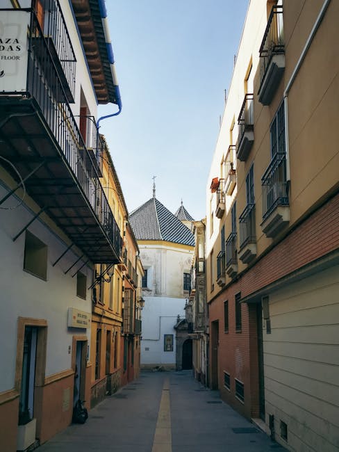 A narrow urban street between residential buildings during daylight, with multi-storey structures on both sides featuring balconies with black metal railings and some with potted plants. The street is paved and contains a yellow line down the center, leading towards a white church building with a conical tiled roof and a central spire visible in the background. The surrounding buildings are constructed with a mix of stucco and brick, with windows, doors, and external fire escapes. Bright natural light illuminates the scene, highlighting the textures of the building facades and the clear blue sky overhead. This setting is typical of a European city, suitable for house removals or furniture transport services provided by Man with Van Southfields, especially during a home relocation or packing and moving process across tight streets.