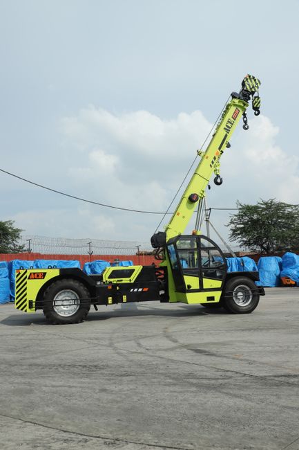 A lime green mobile crane with black and yellow striped warning markings on the rear section is positioned on a paved surface, with its extendable arm fully lifted into the air. The crane's cab is enclosed with glass windows, and there are safety hooks and pulleys attached to the end of the arm. Behind the crane, a row of large, blue protective covers is visible, possibly covering stored equipment or materials, with a chain-link fence and some trees further in the background under a cloudy sky. This scene depicts a typical setup for lifting or transporting heavy objects during a house removal or relocation process, with the crane ready to assist in the safe handling of heavy furniture or appliances, supporting the logistics involved in home moving services provided by companies such as Man with Van Southfields.