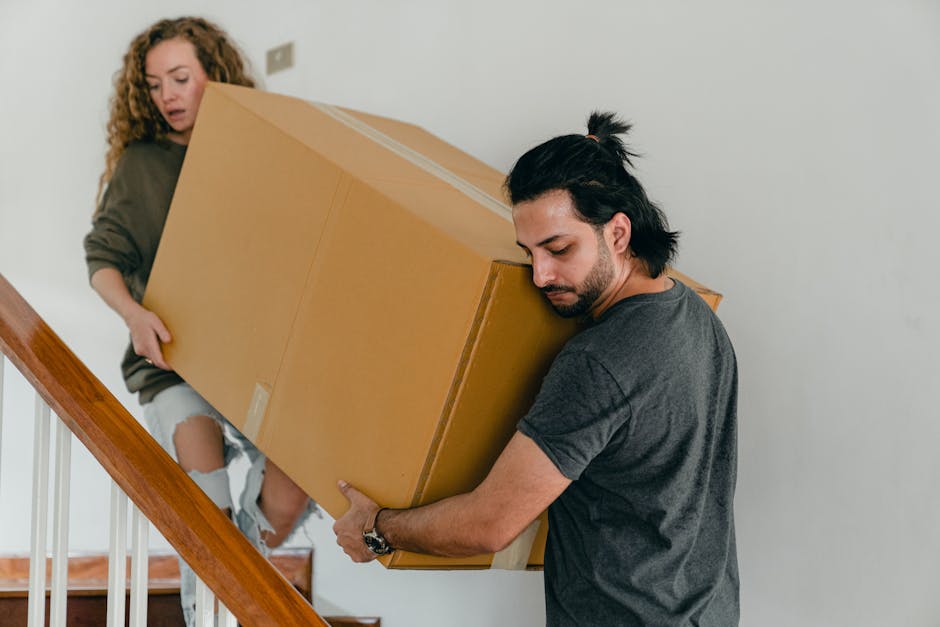 A man with dark hair tied back in a ponytail wearing a dark grey t-shirt is carefully carrying a large cardboard box up a staircase inside a residential property, supported by both hands, with a focused expression. A woman with curly hair, dressed in a grey sweater and ripped jeans, is standing behind him, holding the other side of the box, assisting with the heavy item. The staircase features white balustrades and wooden banisters, and the walls are plain white, illuminated by natural light. The scene captures the process of home relocation or furniture transport, with the pair working together to load the box into the upper floor, indicative of professional removals or moving services by Man with Van Southfields as part of a packing and moving operation.