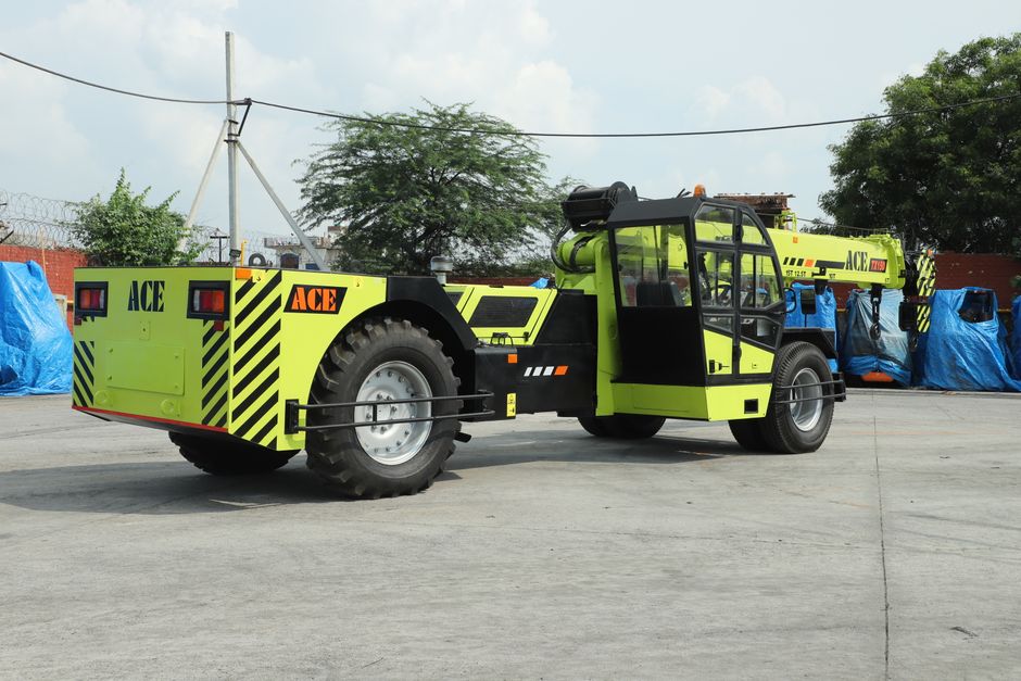 A lime green mobile crane with black and yellow striped warning markings on the rear section is positioned on a paved surface, with its extendable arm fully lifted into the air. The crane's cab is enclosed with glass windows, and there are safety hooks and pulleys attached to the end of the arm. Behind the crane, a row of large, blue protective covers is visible, possibly covering stored equipment or materials, with a chain-link fence and some trees further in the background under a cloudy sky. This scene depicts a typical setup for lifting or transporting heavy objects during a house removal or relocation process, with the crane ready to assist in the safe handling of heavy furniture or appliances, supporting the logistics involved in home moving services provided by companies such as Man with Van Southfields.