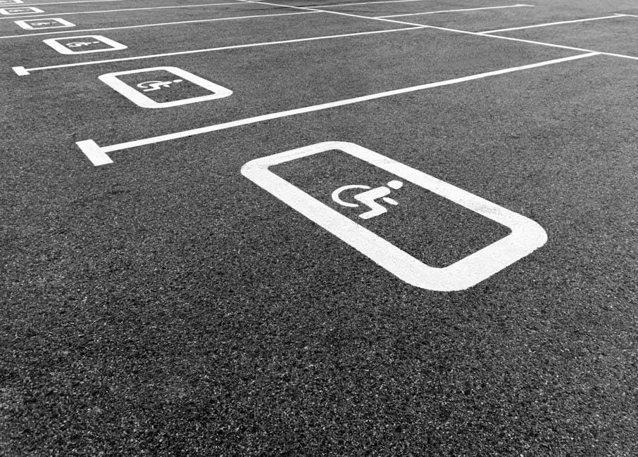 Close-up view of an asphalt parking lot surface with white painted markings indicating designated accessible parking spaces. One marked space prominently displays a wheelchair symbol within a rectangular outline, indicating disabled parking. The markings are clean and sharply defined, with surrounding asphalt showing a textured surface. In the background, additional parking spaces with similar white outlines and symbols are visible, aligned in a row. The scene is illuminated by natural daylight, capturing a well-maintained, organized parking area. This image is relevant to house removals and moving logistics, illustrating accessible parking considerations for loading and unloading during home relocations, which is a service provided by Man with Van Southfields, featured on their SW18 removals guide webpage.