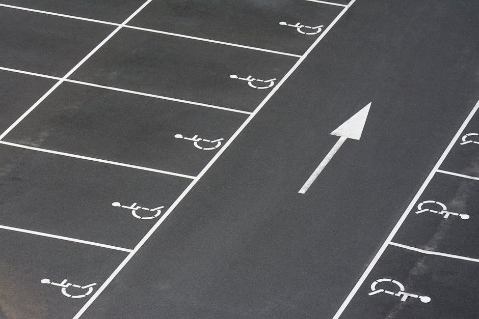 Close-up view of an asphalt parking lot surface with white painted markings indicating designated accessible parking spaces. One marked space prominently displays a wheelchair symbol within a rectangular outline, indicating disabled parking. The markings are clean and sharply defined, with surrounding asphalt showing a textured surface. In the background, additional parking spaces with similar white outlines and symbols are visible, aligned in a row. The scene is illuminated by natural daylight, capturing a well-maintained, organized parking area. This image is relevant to house removals and moving logistics, illustrating accessible parking considerations for loading and unloading during home relocations, which is a service provided by Man with Van Southfields, featured on their SW18 removals guide webpage.