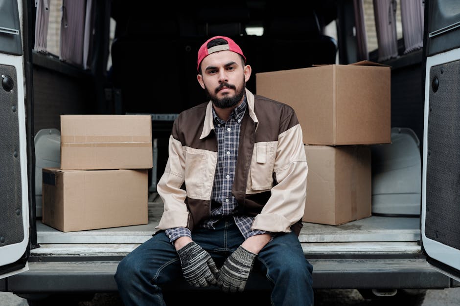 A young man with a serious expression, wearing a black cap and a white polo shirt, is standing inside the open rear of a moving van parked outside a property. Behind him, the van is filled with stacke