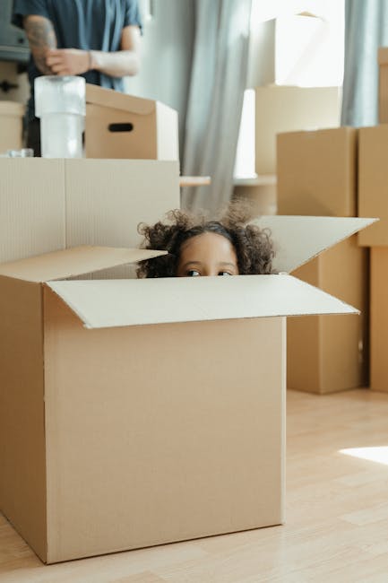 A young child with curly hair peeking out from an open cardboard moving box placed on a light wooden floor inside a room. Surrounding the child are several other sealed cardboard boxes, some stacked and others positioned on the floor, indicating an ongoing home relocation process. In the background, a person wearing a grey shirt is lifting a sealed plastic bottle, with additional boxes and packing materials visible on a table near a window with curtains, allowing natural light to illuminate the space. The scene reflects packing and moving activities typical of a house removals service, with furniture and boxes prepared for transport, as seen in the work carried out by Man with Van Southfields.