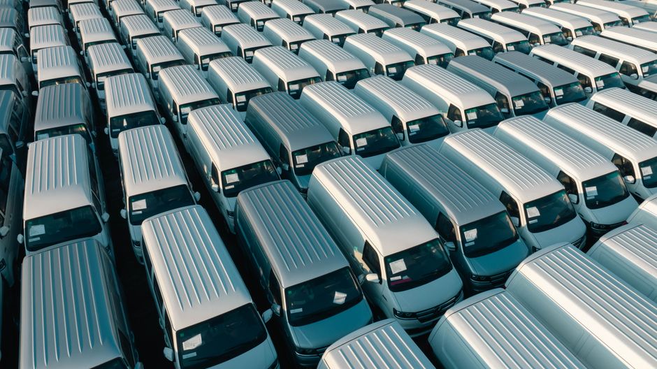 An aerial view shows a large parking lot filled with rows of identical silver vans, neatly arranged with minimal spacing between them. The vans are uniform in size and color, featuring ribbed metal ro