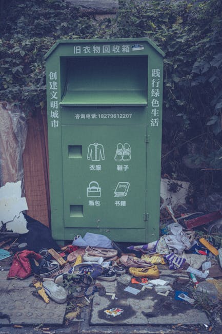 A green old clothing recycling bin situated outdoors next to a wooded area, with scattered items including discarded clothing, shoes, bags, and miscellaneous waste on the ground in front of it. The bin features Chinese characters and icons indicating it is for clothes, shoes, and other textiles, with additional Chinese text promoting recycling and environmental awareness. The scene suggests a collection point for household textiles and clothing scraps, commonly found in urban or suburban environments. Man with Van Southfields may handle the removal and proper disposal of unwanted household items during home relocations or moving services, ensuring waste is responsibly managed. The environment is well-lit, with natural daylight illuminating the clutter and bin, emphasizing the importance of cleaning and waste separation in efficient home and furniture removals and transport logistics.