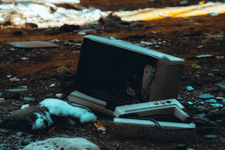An old, discarded computer monitor and keyboard lie on the ground amidst dirt, small rocks, and patches of snow in an outdoor setting. The monitor is tilted backward with visible internal wiring and a dark screen, while the keyboard is partially detached and lying next to it. The surrounding environment includes scattered debris, some plastic waste, and cold weather signs suggesting recent snow. The scene depicts environmental neglect and improper disposal of electronic waste. This image may relate to discussions on waste management, recycling regulations, or community cleanup efforts as part of house or furniture removals, emphasizing proper disposal during moves in Southfields, SW18. Man with Van Southfields is involved in safe and compliant relocation services, ensuring minimal environmental impact during property clearances or move-related waste disposal.