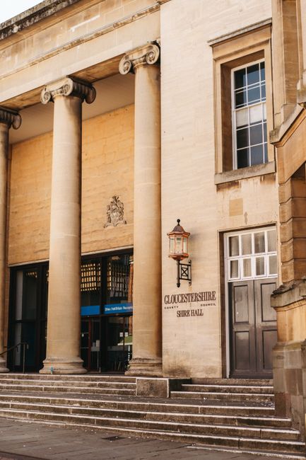 The image shows the exterior of a historic building with classical architectural features, including two large stone columns with ornate capitals supporting a horizontal entablature. The building's façade is made of light-colored stone, with a small inscription sign reading 'GLOUCESTERSHIRE SHIRE HALL' positioned to the right of the entrance. A traditional-style lantern is mounted beside the sign. The entrance area has a set of stone steps leading up to large glass doors, partially visible in the image. To the right of the doors, there is a tall, rectangular window with multiple panes, and nearby wall vents or panels. The scene is well-lit with natural daylight, highlighting the building's detailed stonework. This setting may relate to house removals or relocation services working around notable public buildings, as referenced by the page title concerning Southfields removals and London permits.
