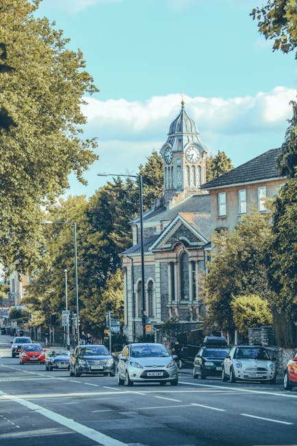 A street scene showing parked cars along the pavement in front of a historic building with a clock tower topped by a pointed spire, situated among large leafy trees under a partly cloudy blue sky. The building appears to be a church or town hall with classic architectural features, including arched windows and decorative brickwork. In the context of house removals, the image captures a typical urban environment where furniture and household items might be loaded into a van for local relocation services, performed by Man with Van Southfields. Visible street furniture includes streetlights and traffic signs, indicating a busy residential and commercial area. The scene reflects a typical setting for home relocation activities, with the environment suitable for moving logistics and transport of packed boxes or large furniture items, supported by professional removal services in Southfields.
