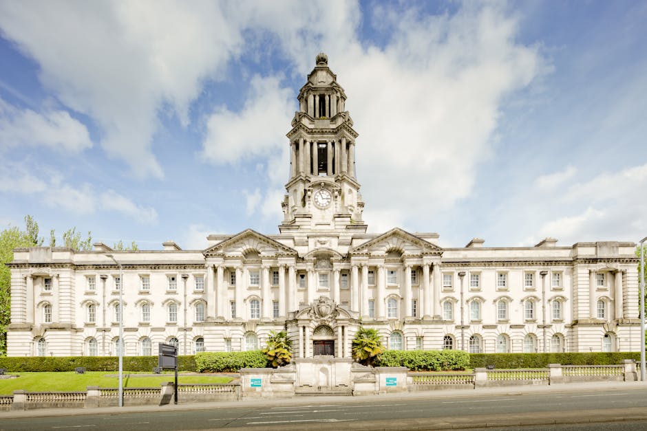 The image shows the exterior of a historic building with classical architectural features, including two large stone columns with ornate capitals supporting a horizontal entablature. The building's façade is made of light-colored stone, with a small inscription sign reading 'GLOUCESTERSHIRE SHIRE HALL' positioned to the right of the entrance. A traditional-style lantern is mounted beside the sign. The entrance area has a set of stone steps leading up to large glass doors, partially visible in the image. To the right of the doors, there is a tall, rectangular window with multiple panes, and nearby wall vents or panels. The scene is well-lit with natural daylight, highlighting the building's detailed stonework. This setting may relate to house removals or relocation services working around notable public buildings, as referenced by the page title concerning Southfields removals and London permits.