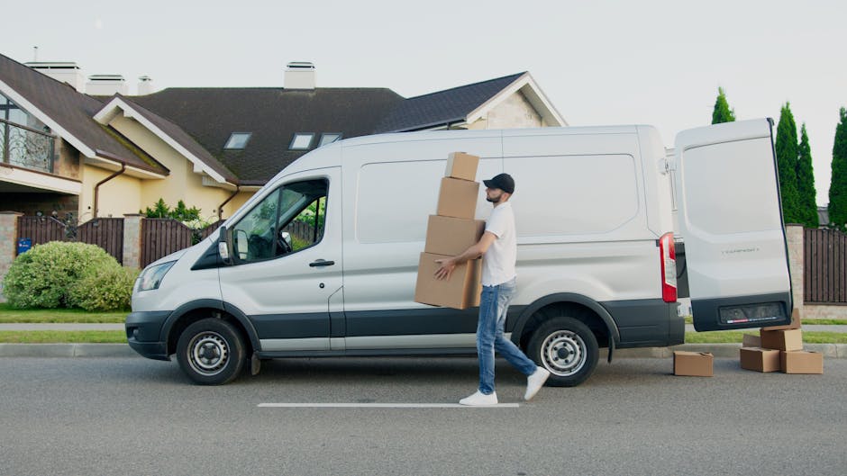 A man wearing a white t-shirt, blue jeans, white sneakers, and a black cap is seen standing beside a white panel van parked on a residential street. He is carrying three cardboard boxes stacked togeth
