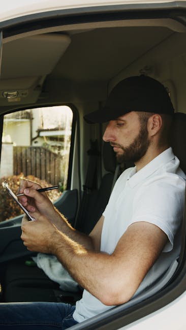 A man wearing a white t-shirt, blue jeans, white sneakers, and a black cap is seen standing beside a white panel van parked on a residential street. He is carrying three cardboard boxes stacked togeth